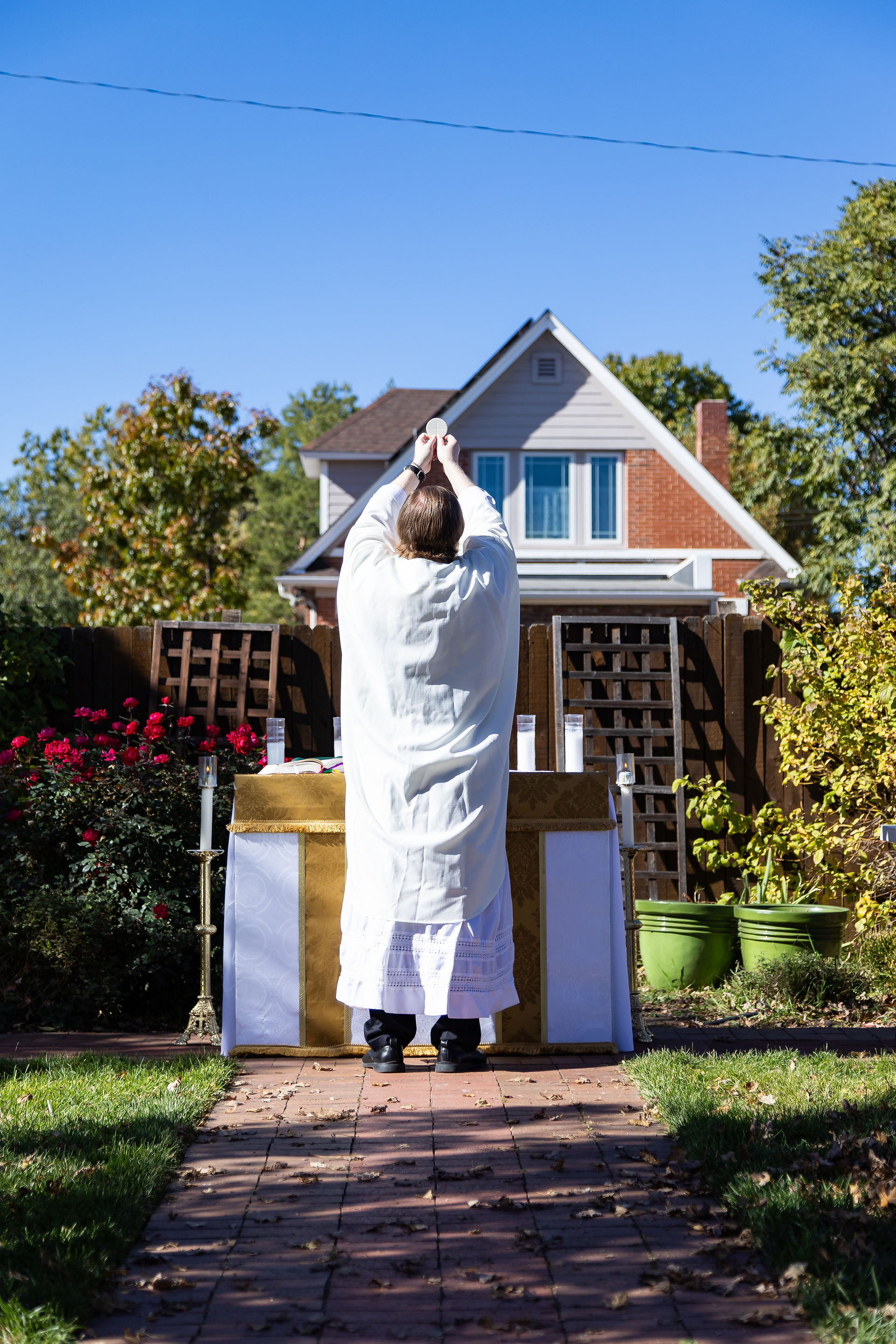 Clergy in white robe holds up a chalice outdoors. Background shows a house, red roses, and green plants under a clear blue sky.