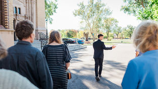 A group follows a man in a suit pointing ahead, near a church with ornate architecture. Trees and parked cars in the sunny background.