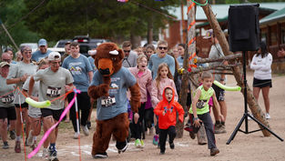 Runners, including a person in a buffalo costume, race outdoors on a dirt path, surrounded by spectators. Energetic atmosphere, colorful attire.