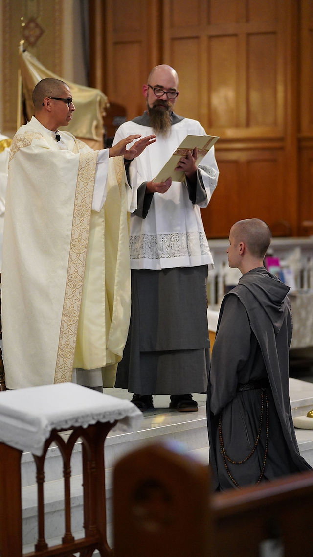 A priest in a white robe gestures while another man holds a book. A kneeling person in a gray robe listens. Wooden panels in background.