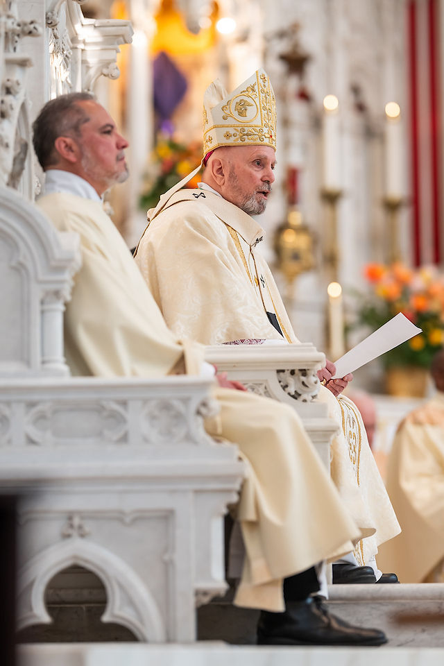 Two clergy in ornate vestments sit on a white stone chair in a church, holding papers. Candles and flowers in the background.