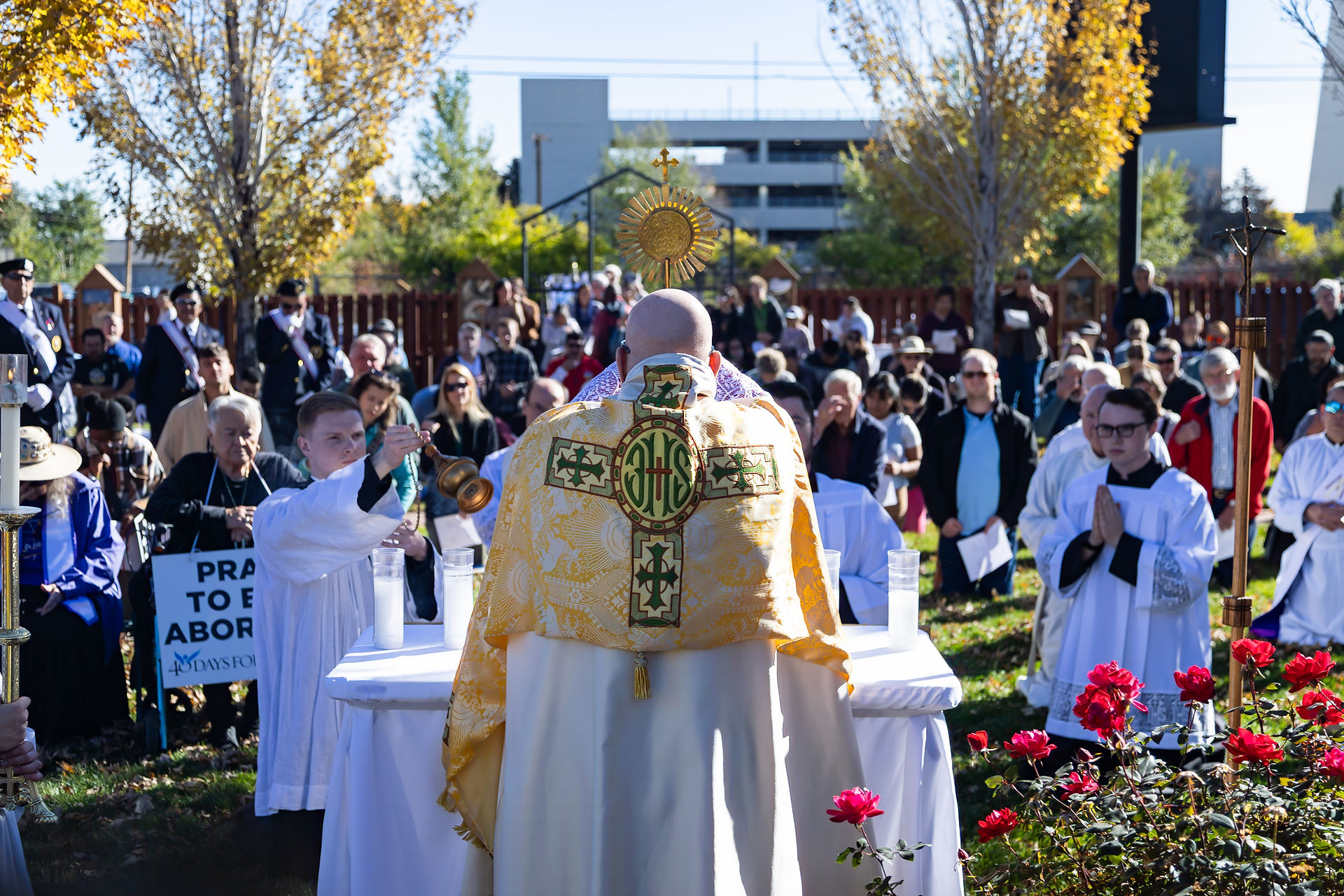 Archbishop in ornate vestment leads outdoor religious ceremony; crowd watches. Roses in foreground, fall trees in background. Sign reads "Pray to End Abortion."