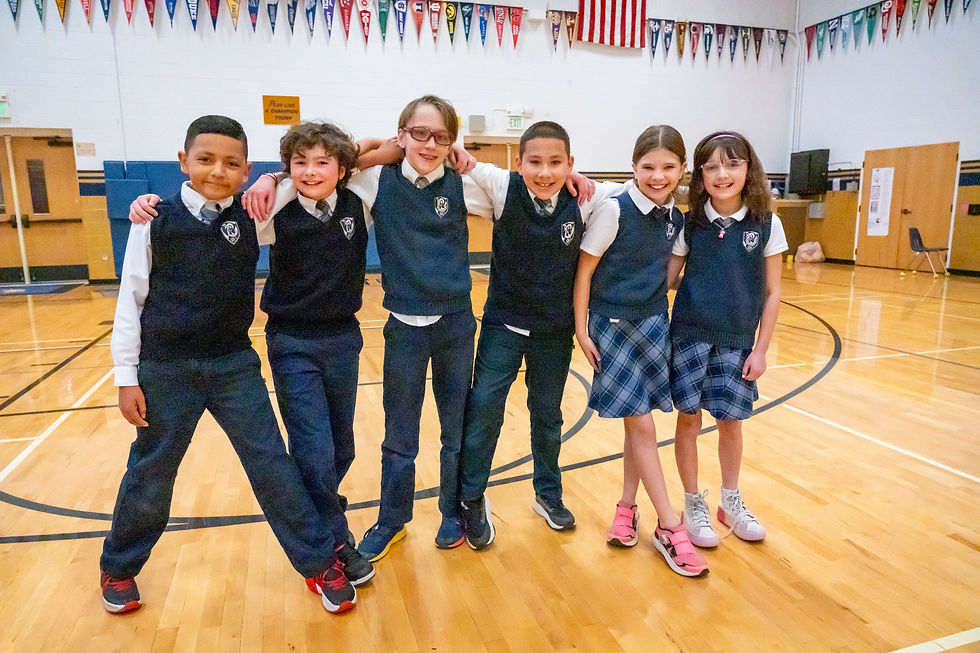 "Six children in school uniforms stand arm-in-arm, smiling in a gymnasium with a wooden floor and basketball court markings. Flags hang on the walls, including a prominently displayed American flag.