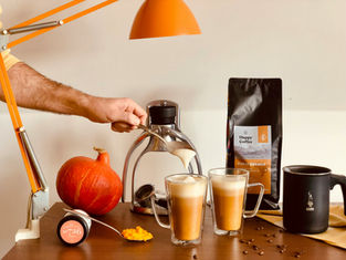 Arm pours milk into coffee glasses on a table with a pumpkin, coffee pods, "Happy Coffee" bag, and orange lamp. Cozy and autumnal mood.