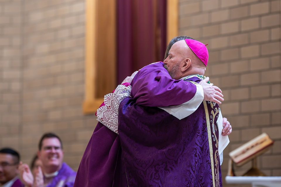 Two clergy in purple robes warmly embrace inside a brick church. Others in the background clap and smile, creating a joyful atmosphere.
