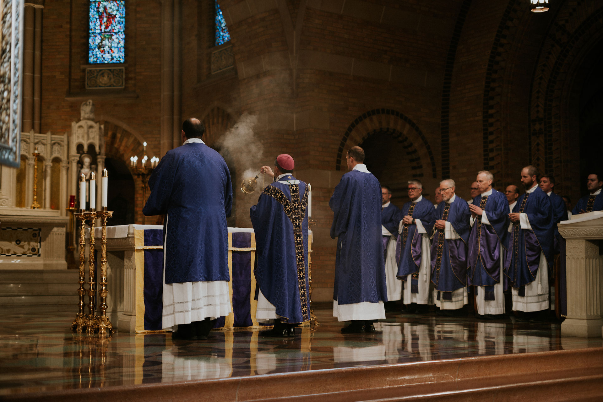 Clergy in purple robes perform a ritual in a church with stained glass and candles. One holds incense, creating a solemn atmosphere.