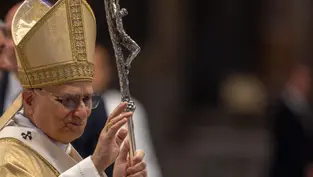 A religious leader in ornate gold vestments and hat waves, holding a silver staff with a cross. The background is blurred.