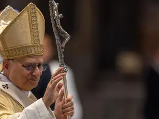 A religious leader in ornate gold vestments and hat waves, holding a silver staff with a cross. The background is blurred.