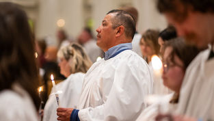 People in white gowns hold lit candles in a solemn ceremony. The setting is bright and blurred, evoking a peaceful, reverent mood.