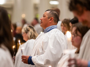 People in white gowns hold lit candles in a solemn ceremony. The setting is bright and blurred, evoking a peaceful, reverent mood.