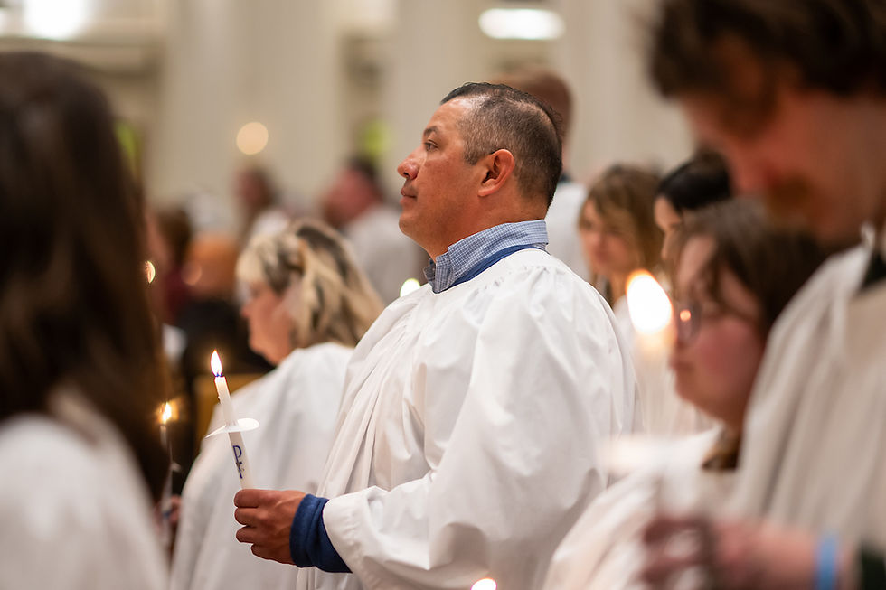 People in white gowns hold lit candles in a solemn ceremony. The setting is bright and blurred, evoking a peaceful, reverent mood.