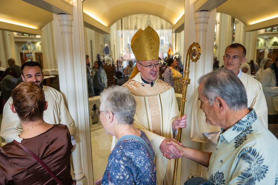Bishop in ornate mitre greets people in a church, holding a gold staff. Warm, friendly setting with mirrored background.