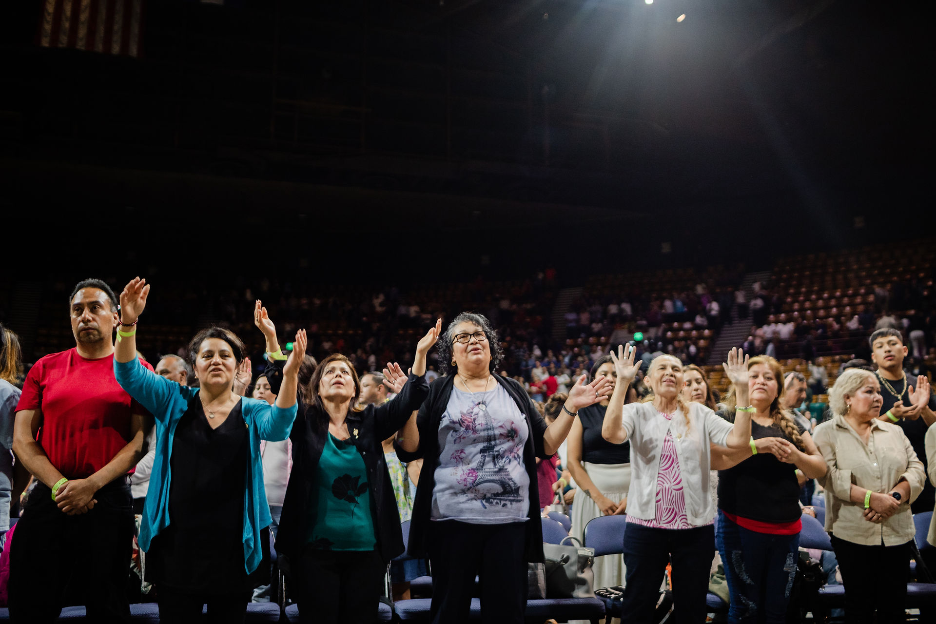 A diverse group of people stands in an auditorium, raising their hands in worship. Dim lighting and a crowded background set a contemplative mood.