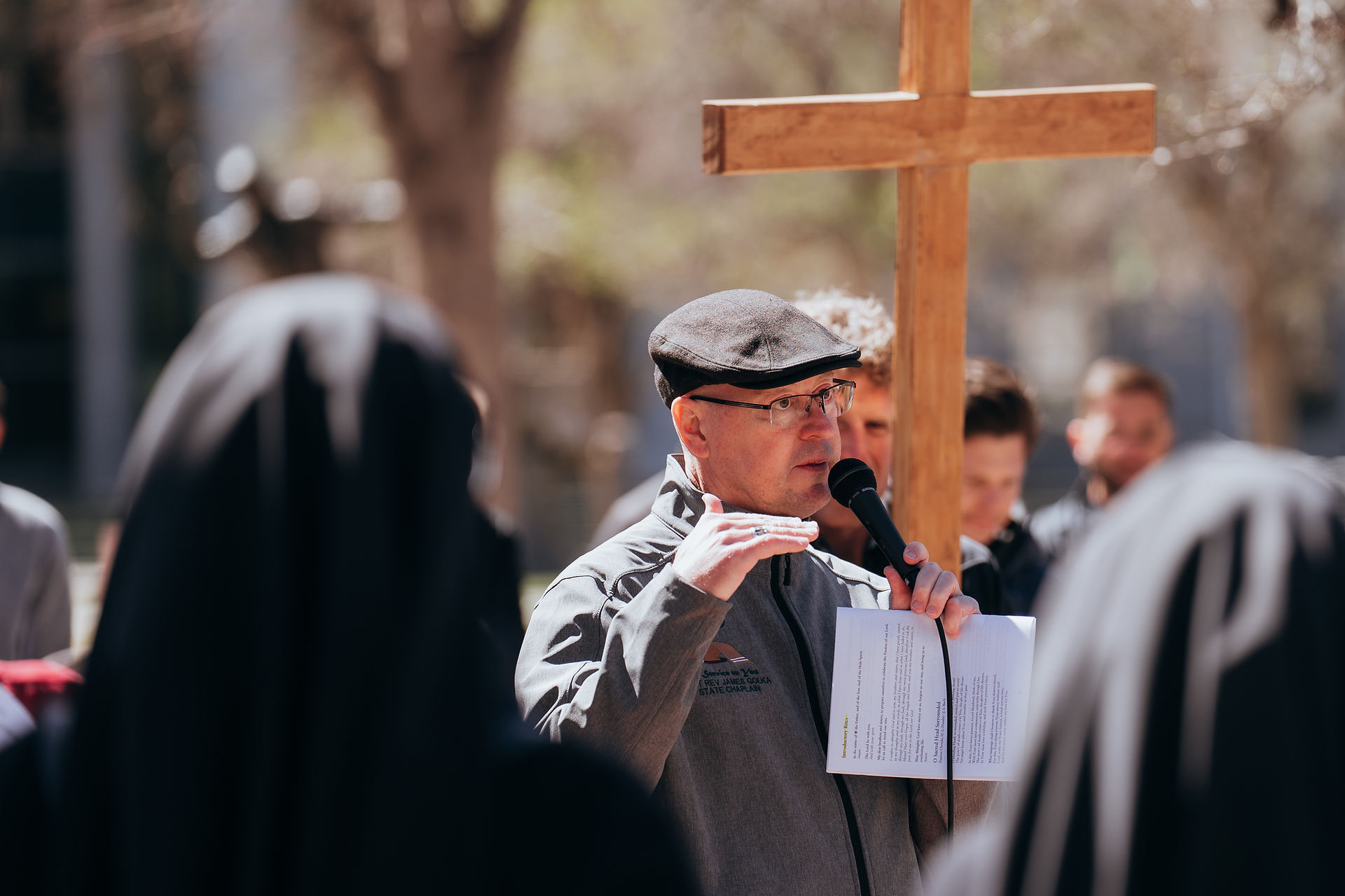 A man speaks into a microphone while holding a paper, standing beside a wooden cross during an outdoor prayer gathering.