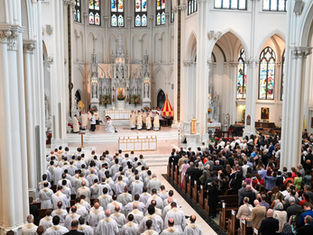 Clergy in white robes lead a ceremony in a grand cathedral with stained glass windows. A large congregation watches solemnly.