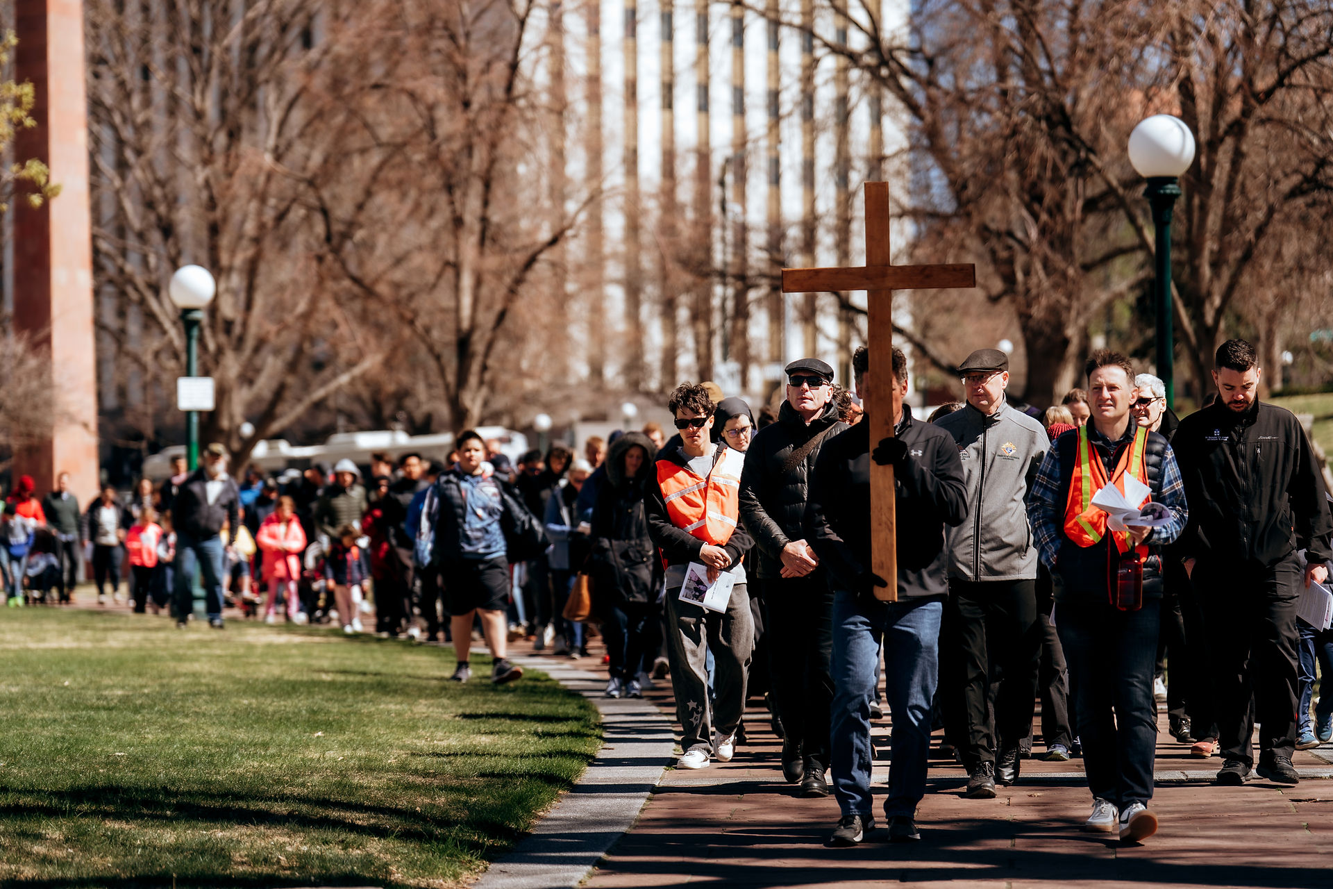 A large group processes along a park path, led by a man carrying a wooden cross during a Way of the Cross devotion.