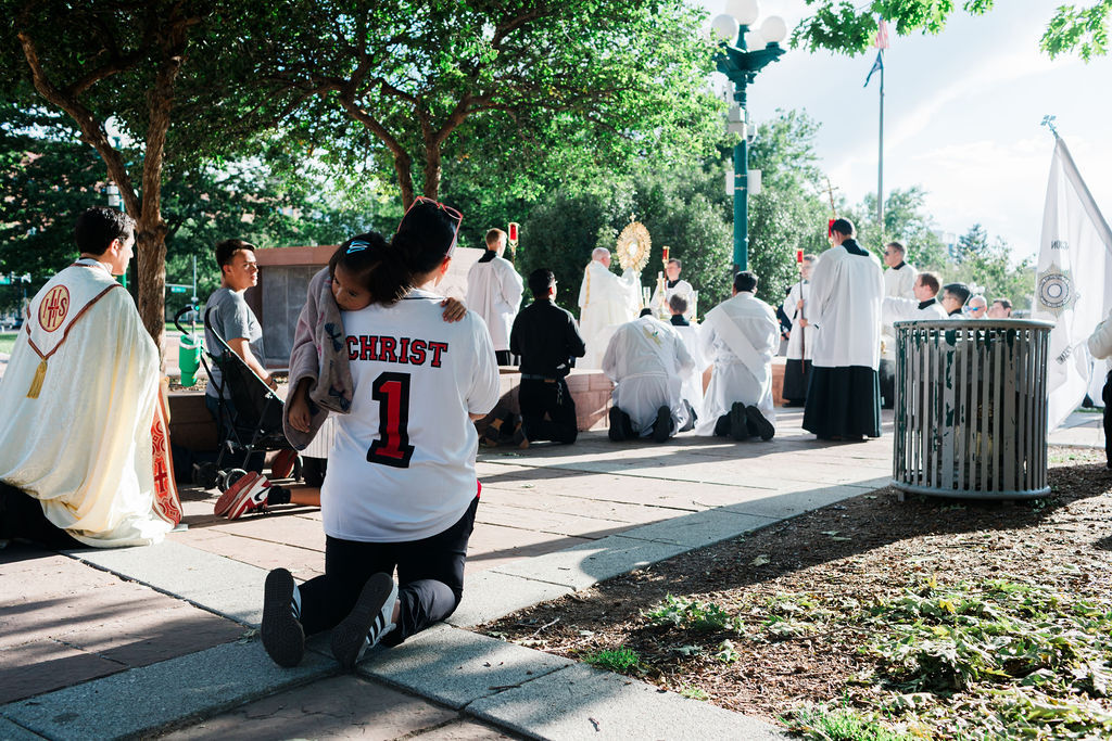 Outdoor Catholic service with kneeling clergy and worshippers; woman in CHRIST 1 jersey holds a child under trees.