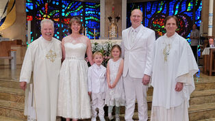 A family and two clergy in white attire smile inside a church with colorful stained-glass windows. The mood is joyful and celebratory.