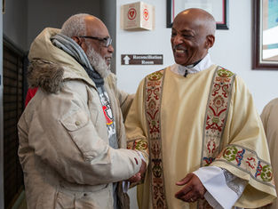 Two men smiling and shaking hands in a church. One wears a beige jacket, the other a patterned robe. Signs indicate a Reconciliation Room.