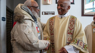 Two men smiling and shaking hands in a church. One wears a beige jacket, the other a patterned robe. Signs indicate a Reconciliation Room.
