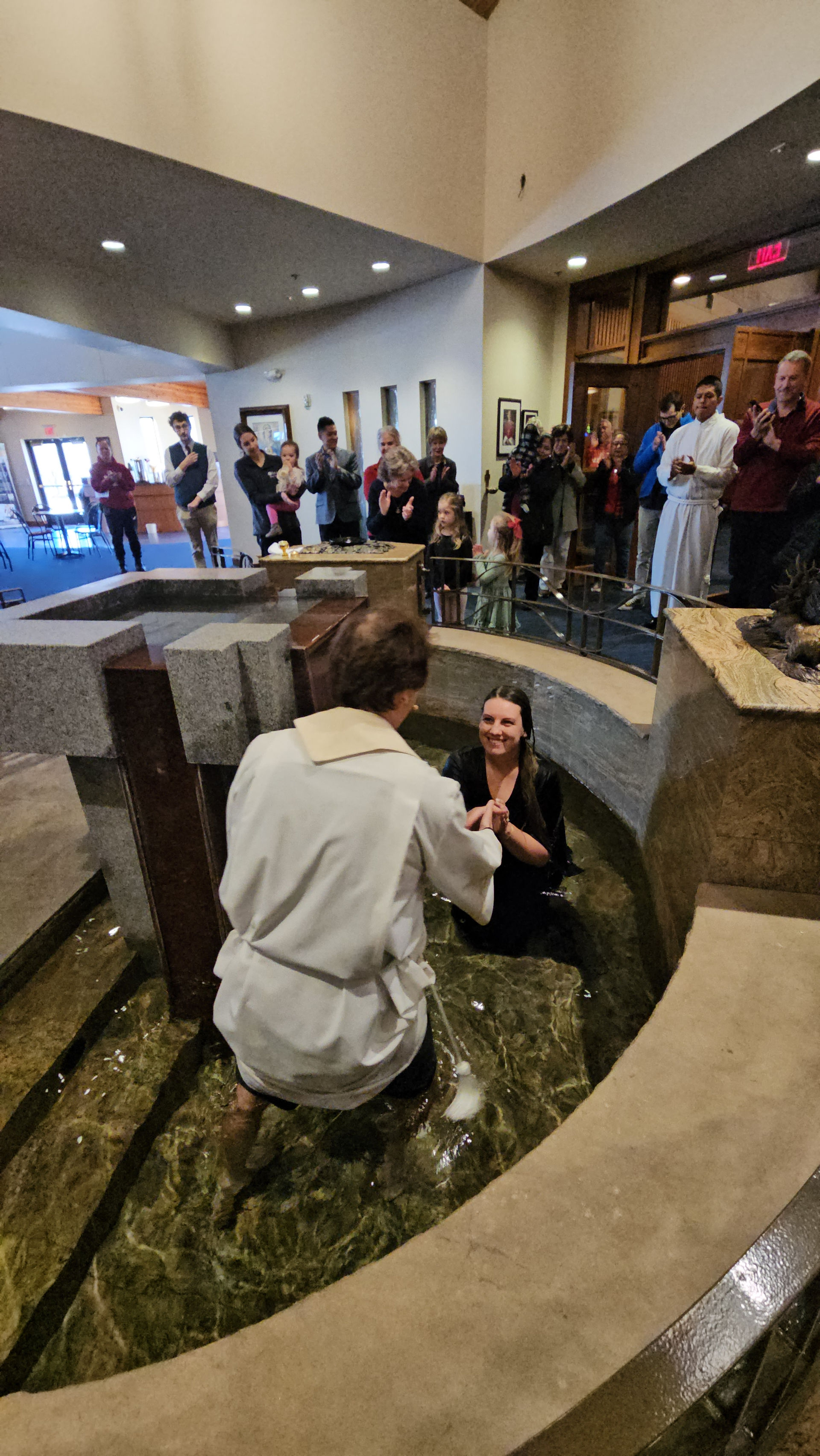 A person in white baptizes a smiling woman in a stone font, surrounded by onlookers clapping in a warmly lit church interior.