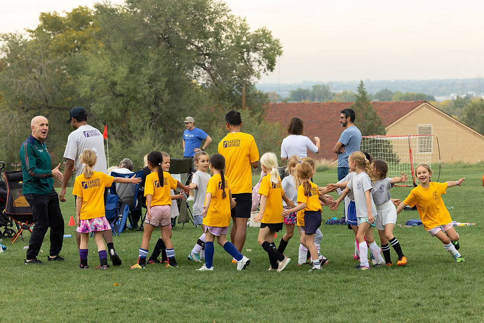 Kids in yellow and white University Catholic soccer shirts gather on a grassy field with coaches and parents near a goal.