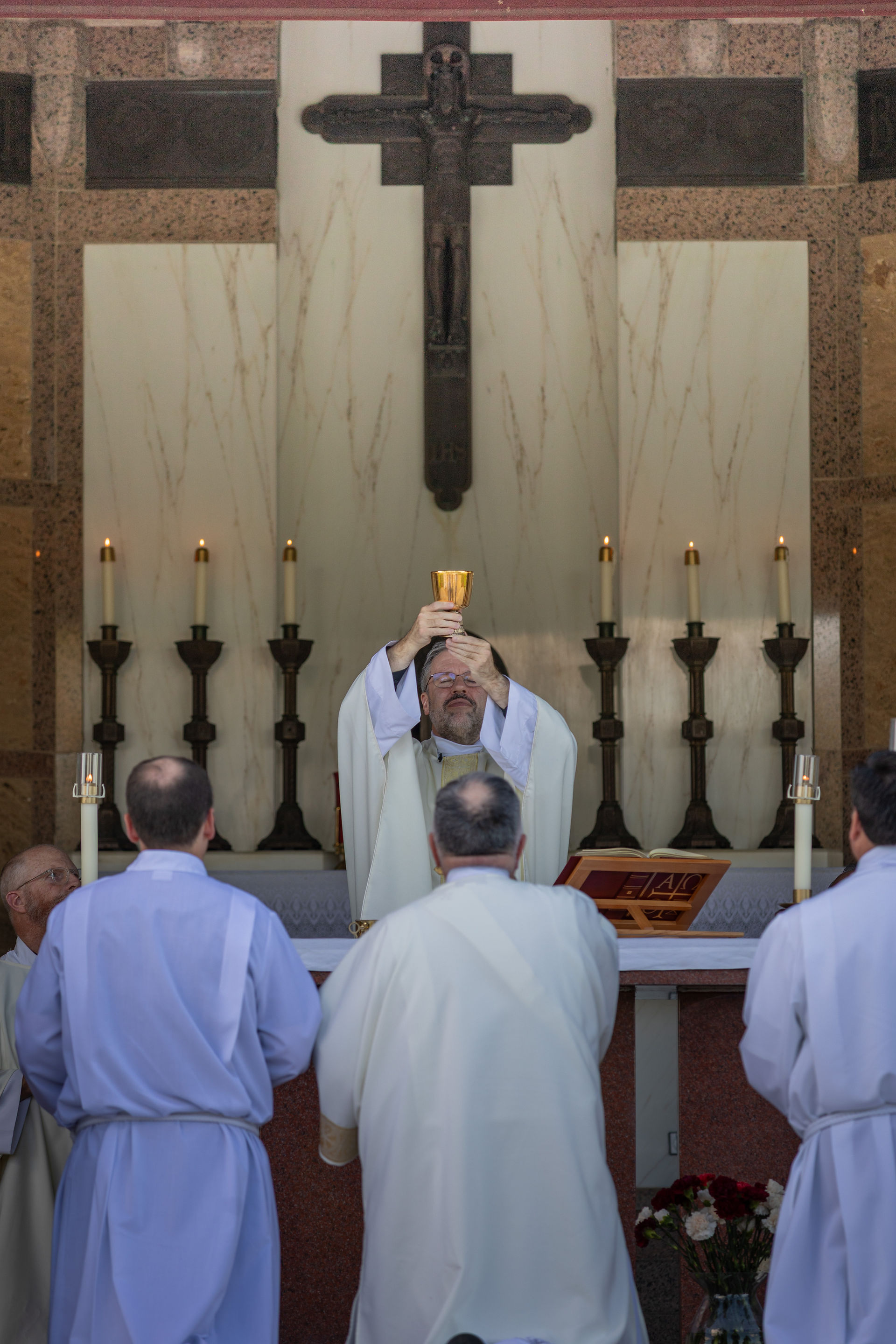 Priest holds chalice up during a ceremony in a church. Candles and crucifix in the background. Clergy in white robes observe solemnly.