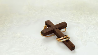 Wooden cross tied with twine, two gold rings on it, set on delicate white lace. Symbolic, peaceful atmosphere.