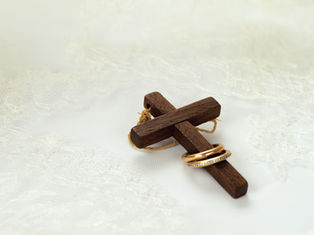 Wooden cross tied with twine, two gold rings on it, set on delicate white lace. Symbolic, peaceful atmosphere.