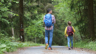 Father and daughter with backpacks walking on a forest trail. Lush green trees surround them, creating a peaceful vibe.