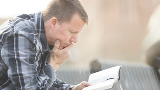 A man in a plaid shirt reads thoughtfully outdoors on a sunny day, holding a book with focus and concentration.