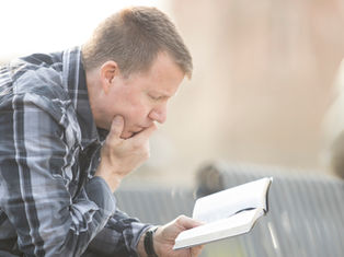 A man in a plaid shirt reads thoughtfully outdoors on a sunny day, holding a book with focus and concentration.