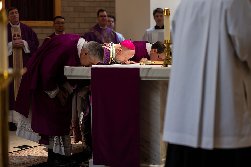 Clergy in purple robes bow at an altar in a church setting, candles and other staff in background, creating a solemn atmosphere.