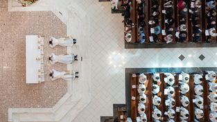 Aerial view of three priests lying prostrate before an altar in a church, surrounded by seated congregation, highlighting ceremonial solemnity.