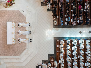 Aerial view of three priests lying prostrate before an altar in a church, surrounded by seated congregation, highlighting ceremonial solemnity.