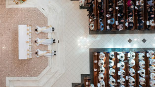 Aerial view of three priests lying prostrate before an altar in a church, surrounded by seated congregation, highlighting ceremonial solemnity.
