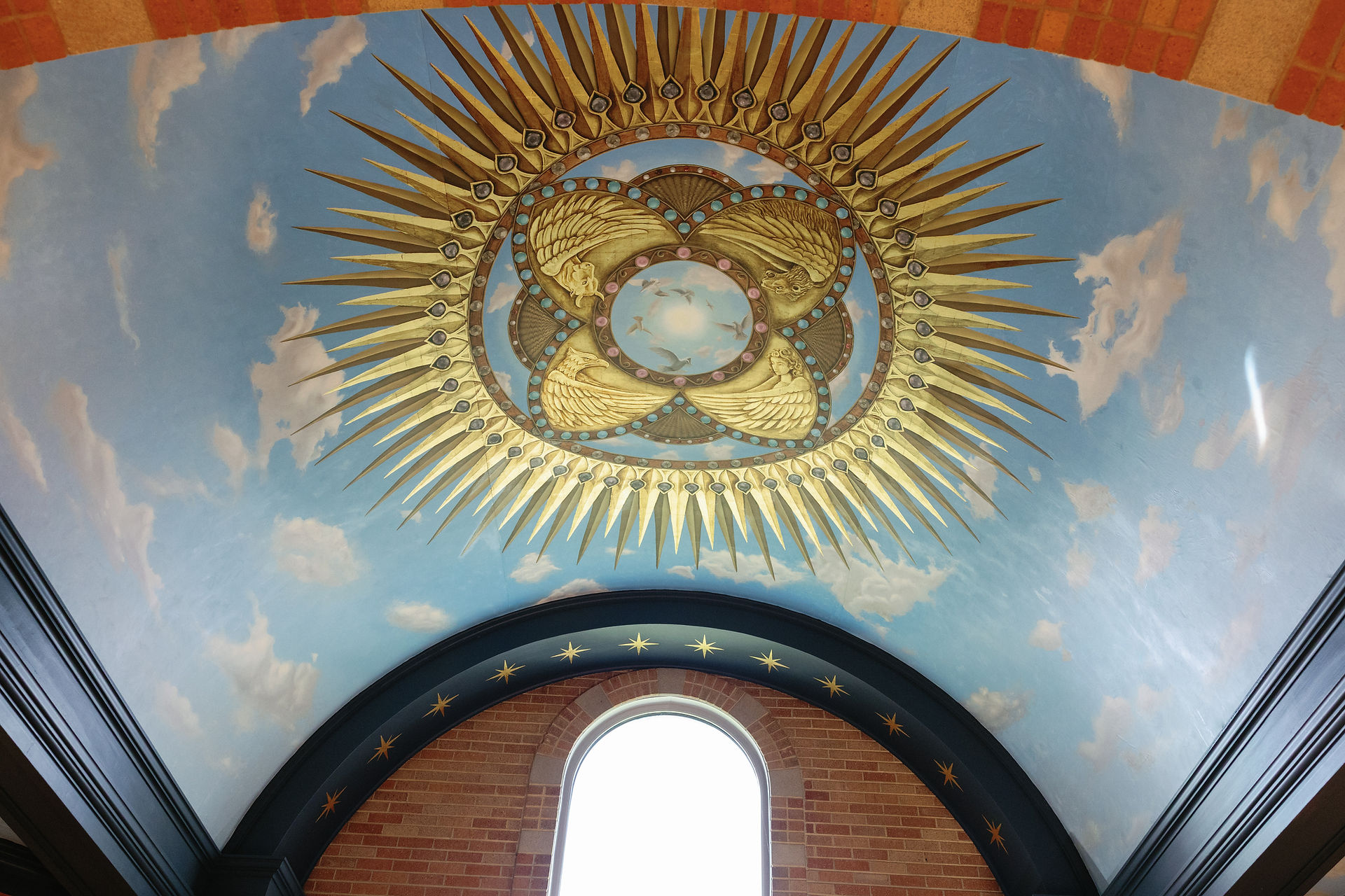 Ornate ceiling art with golden sunburst, angels, and blue sky background. Brick arch window below, creating a serene atmosphere.