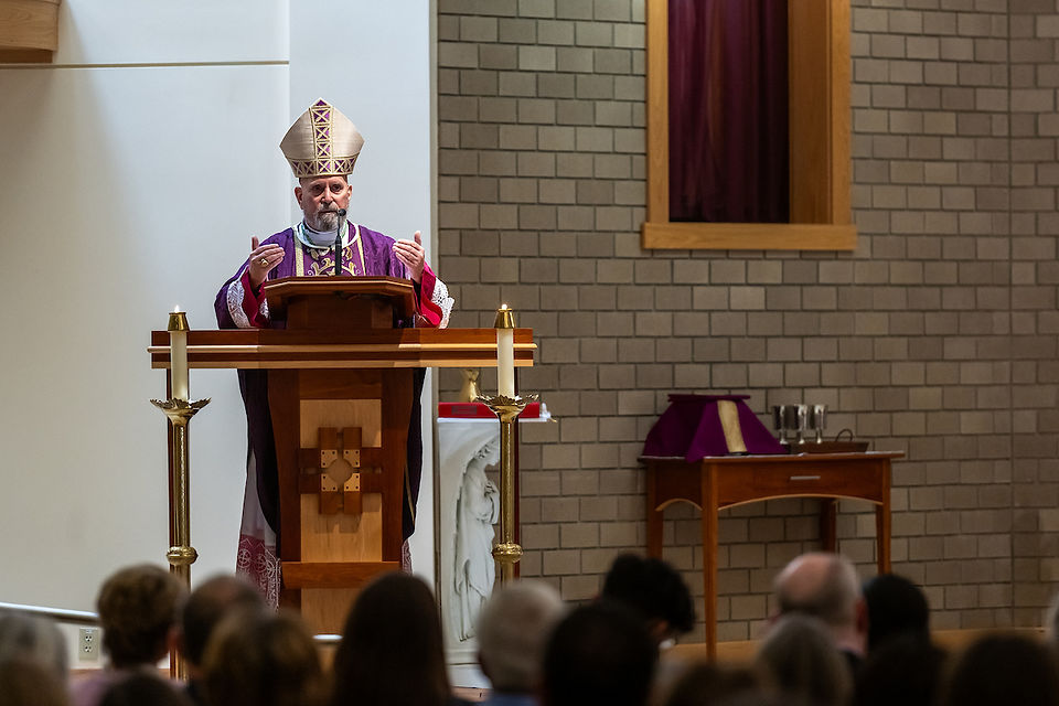 Bishop in ornate vestments speaks at pulpit in church, flanked by candles. Congregation listens. Brick wall and purple draped table in view.