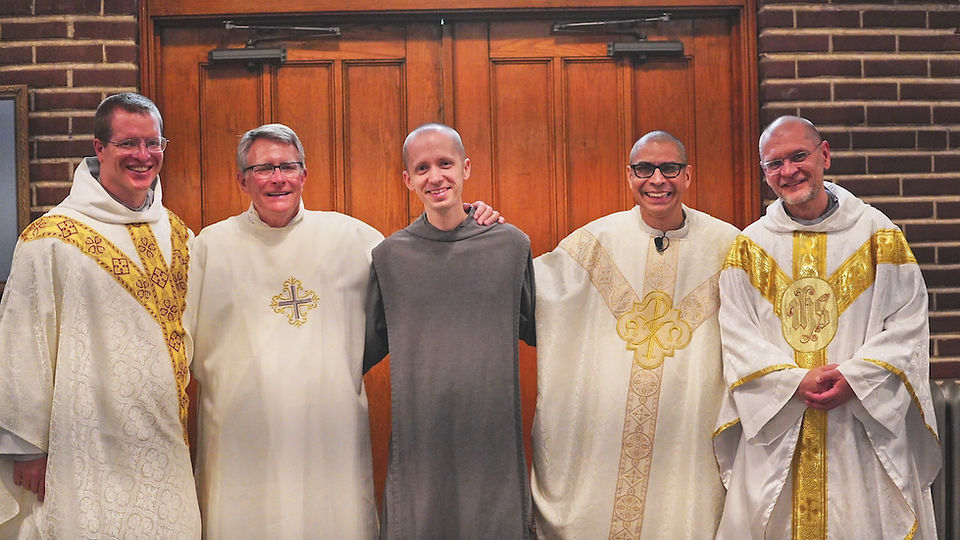 Five men in religious robes smiling in front of wooden doors. Robes are white with gold details. Brick wall visible in background.