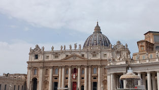 Crowd gathers in St. Peter's Square, Vatican City, under a cloudy sky, with St. Peter's Basilica and a fountain visible in the background.