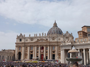 Crowd gathers in St. Peter's Square, Vatican City, under a cloudy sky, with St. Peter's Basilica and a fountain visible in the background.
