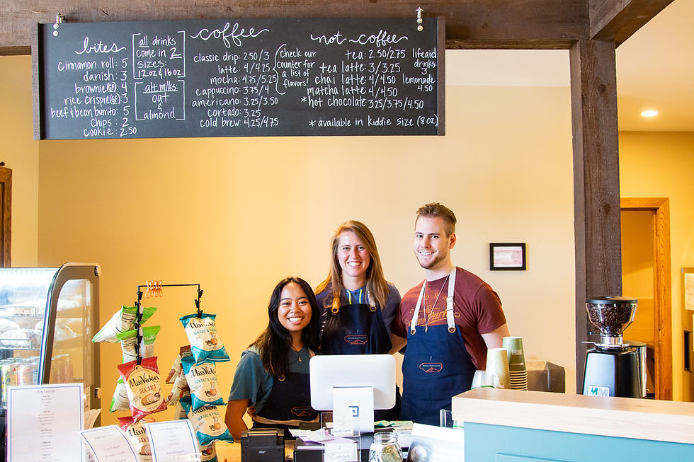 Three smiling baristas behind a coffee shop counter with a menu board above. Display of chips, a payment terminal, and a minimalist decor.