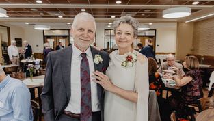 Elderly couple smiling at a gathering, dressed formally with floral corsages. Bright room with people socializing in the background.