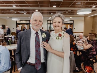 Elderly couple smiling at a gathering, dressed formally with floral corsages. Bright room with people socializing in the background.