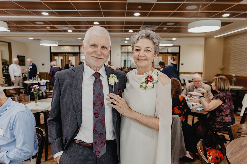 Elderly couple smiling at a gathering, dressed formally with floral corsages. Bright room with people socializing in the background.