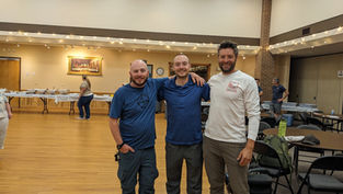 Three smiling men in casual clothes pose in a hall with wooden floors. Tables in the background have food trays. Warm lighting and a relaxed mood.