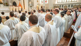 Priests in white robes with cream collars standing in a church. Stained glass windows and altar visible. Formal, solemn atmosphere.