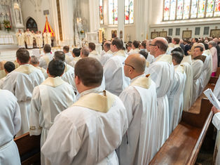 Priests in white robes with cream collars standing in a church. Stained glass windows and altar visible. Formal, solemn atmosphere.