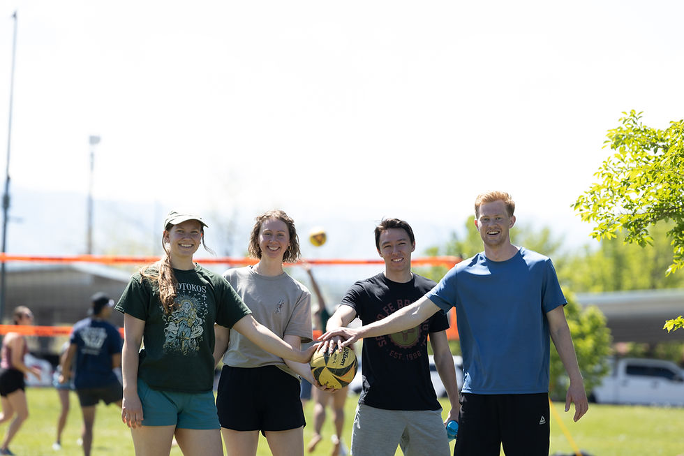 Four people smiling and touching a yellow volleyball outdoors. Background of others playing, grass field, and trees. Sunny day.