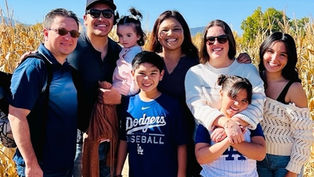 A group of smiling people, including kids, pose outdoors in a cornfield. A boy wears a blue Dodgers T-shirt. Bright, sunny day, clear sky.
